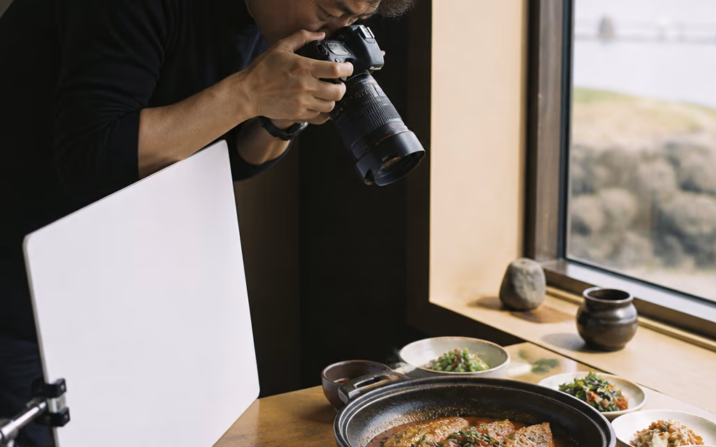 제주 식당 창가 테이블에서 광고 사진가가 자연광 아래 갈치조림 한 상을 카메라로 촬영하는 장면